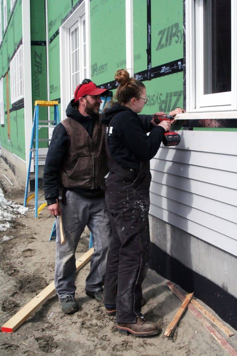 A man watching a woman use a cordless drill on a piece of trim on a house under construction